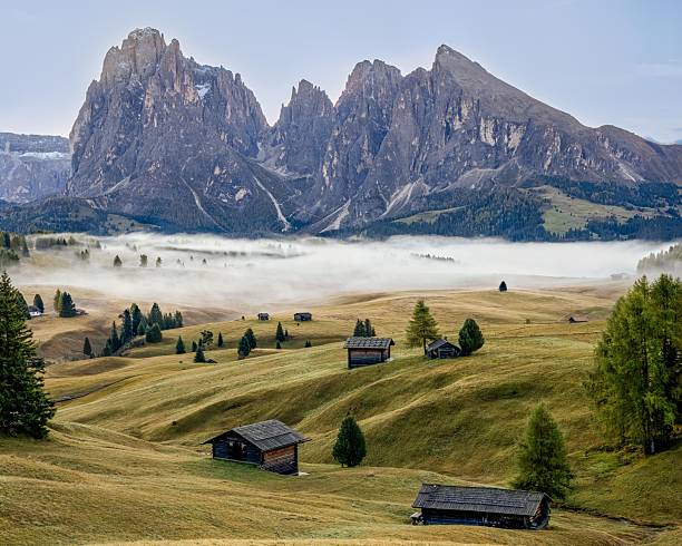 A stunning view of the Dolomites with mist over rolling green hills and scattered wooden huts in Alpe di Siusi, Italy.