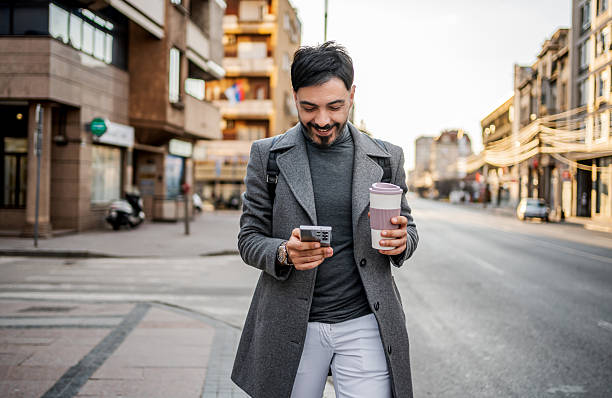 istock Man smiling while checking phone and holding coffee in city 2241242545