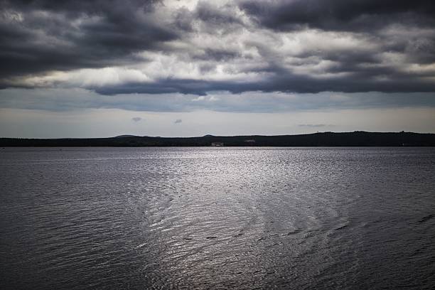 Dramatic stormy sky looms over serene lake with distant trees in a tranquil, melancholic landscape.