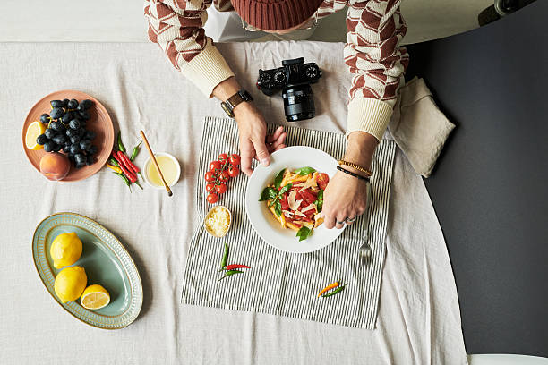 Man arranging food on plate at table, digital camera placed nearby, fresh fruits and vegetables surrounding, hands carefully styling meal for photography