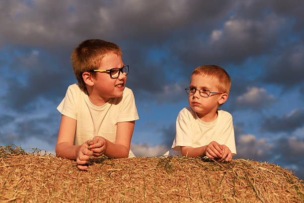Two boys lie on a hay bale in golden sunset light under a picturesque sky and talk to each other. Concept for friendship, childhood, copy space background.