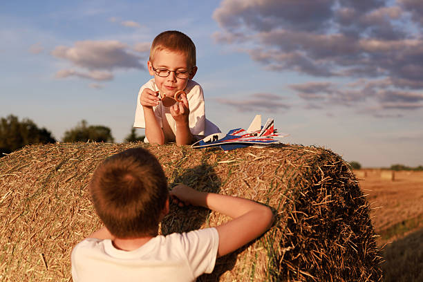 Two boys play on a hay bale with a toy airplane in golden sunset light. Concept for travel, adventure, rural copy space background.