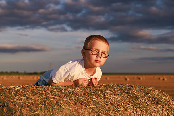 A sad boy with glasses lies on a hay bale in golden sunset light under a picturesque sky. Concept for emotion, childhood, copy space background.