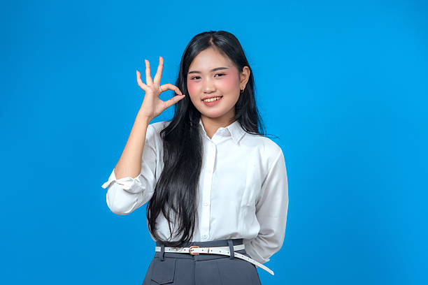 istock Smiling young Asian woman making an OK hand gesture while standing confidently in a white shirt and gray pants against a solid blue background, perfect for advertising and marketing use. 2241268875
