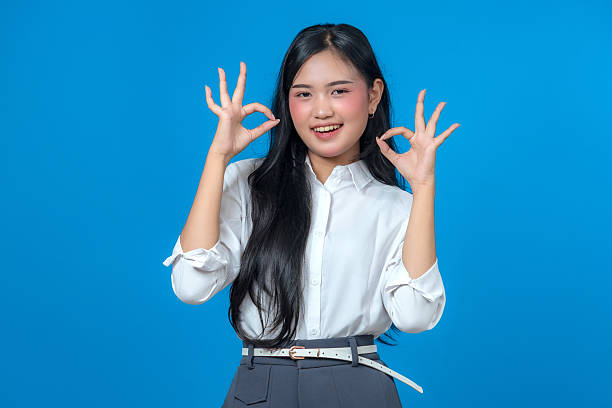 istock Smiling young Asian woman showing double OK hand gestures with both hands, wearing a white shirt and gray pants, standing confidently against a vibrant blue background in studio light. 2241268889