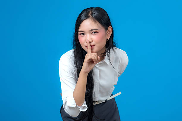 istock Young Asian woman making a shushing gesture with index finger on lips, smiling slightly and leaning forward, wearing white shirt against blue background in a studio shot. 2241269057