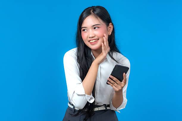 istock Charming young Asian woman in white shirt holding smartphone and smiling while making a playful talking gesture with her hand, standing against a blue background in studio light. 2241269802