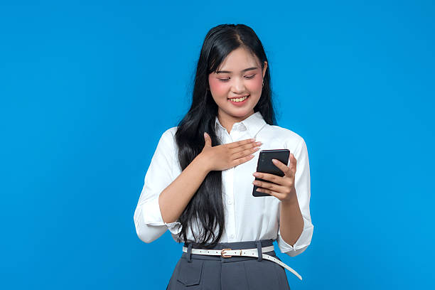 istock Smiling young Asian woman looking at her smartphone while placing her hand on her chest, expressing gratitude or happiness. She stands against a plain blue background in casual attire. 2241269841
