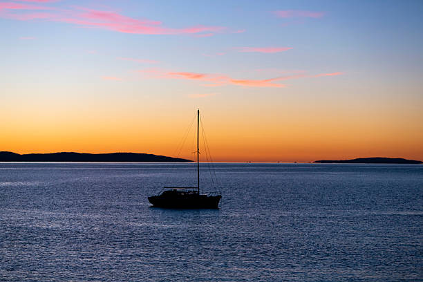 Serene view of a lone sailboat drifting on calm waters under a vibrant sunset sky. The horizon glows with warm orange and pink tones, framed by distant landmasses and a peaceful atmosphere. Ideal for visuals focused on solitude, natural beauty, maritime lifestyle, and scenic ocean moments. No specific location indicated.