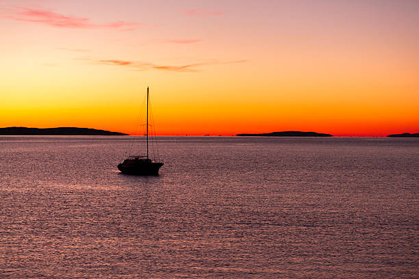 Serene view of a lone sailboat drifting on calm waters under a vibrant sunset sky. The horizon glows with warm orange and pink tones, framed by distant landmasses and a peaceful atmosphere. Ideal for visuals focused on solitude, natural beauty, maritime lifestyle, and scenic ocean moments. No specific location indicated.