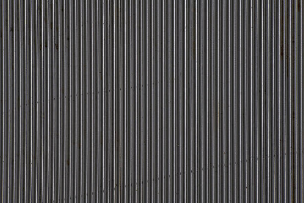 Close-up horizontal texture shot of corrugated metal showing narrow vertical ridges and valleys in a dull grey tone, with faint traces of rust and grime adding subtle wear.