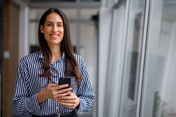 istock Young businesswoman smiling, holding smartphone, and looking at camera while standing in a bright modern office 2241277721