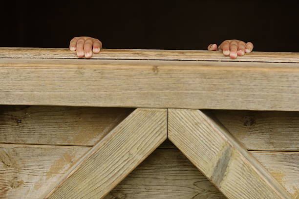A wooden structure with two hands peeking out from behind it. Scene is mysterious and curious