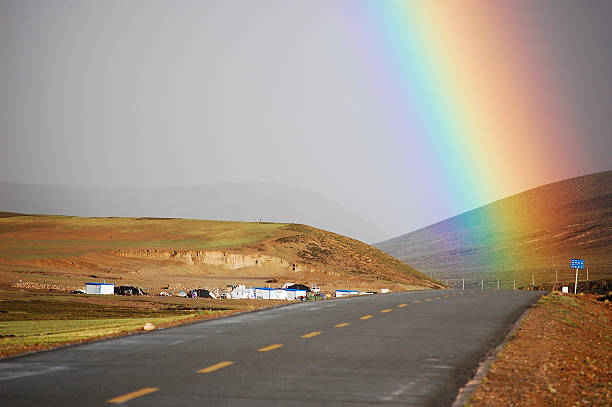 A beautiful rainbow spans over a rural road with rolling hills in the background, showcasing a natural and picturesque scene.