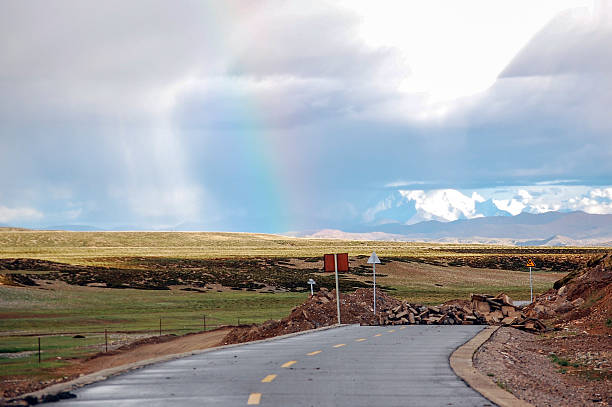 A beautiful rainbow is visible over a paved road in a vast, open landscape with clouds and green fields.