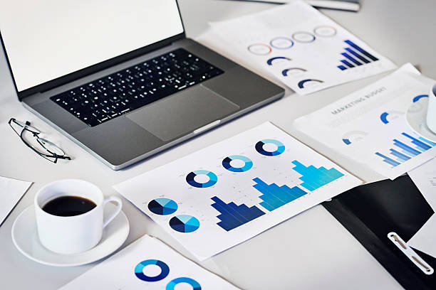 An overhead shot of a clean white desk featuring a laptop, a cup of coffee, reading glasses, and multiple printed business documents displaying various blue-toned charts and graphs (marketing budget, bar charts, pie charts).
