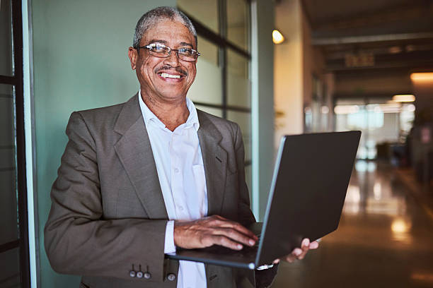 istock Happy Senior Black Businessman Smiling While Using Laptop in Office Hallway 2241294392