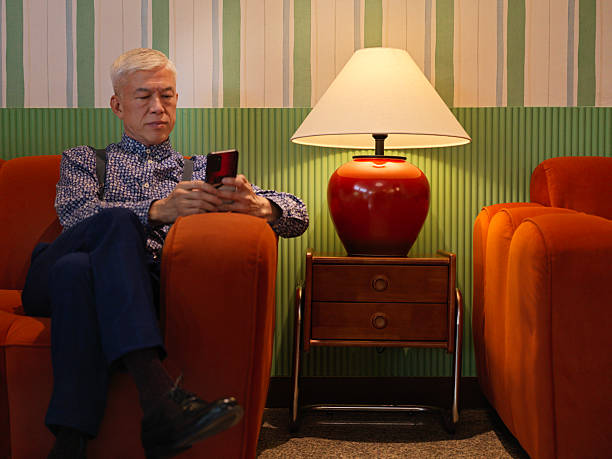 chinese senior man sitting comfortably on a sofa at home, holding and using a smartphone, representing digital lifestyle and modern technology adoption, Kuala Lumpur.