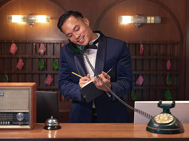 istock Smiling Asian Chinese male Hotel concierge taking notes while talking on the phone at vintage reception counter, luxury hospitality concept 2241306684