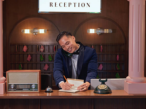 Asian male concierge in a tuxedo multitasking at a vintage-style hotel reception desk, writing notes while speaking on the telephone, representing luxury service, professionalism, and hospitality. Kuala Lumpur.