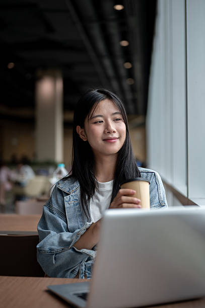 Smiling young asian woman holding coffee while sitting with laptop on table in the cafe or lounge.