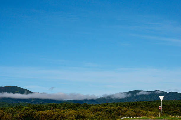 A forested mountain range with drifting mist clouds rises under a blue sky, while a road sign stands in the foreground near grassy fields. Nature of Sakhalin Island.