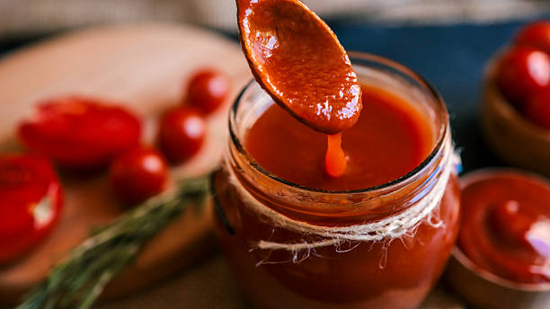A close-up shot captures the rich, red tomato sauce being poured from a wooden spoon into a glass jar, with fresh tomatoes in the background.
