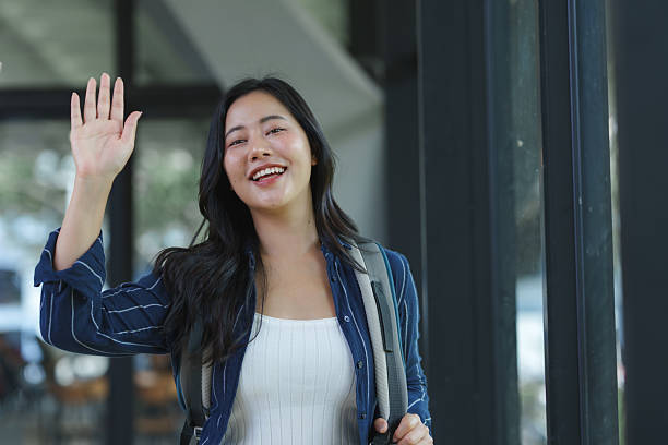 Happy young woman traveler with backpack smiling and waving hand in greeting or farewell, cheerful college student looking at camera
