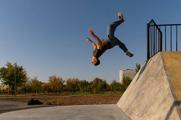 A male freerunner performs a backflip off a sloped concrete wall in an outdoor park, captured midair with extended limbs under clear skies, showcasing athletic precision and agility.