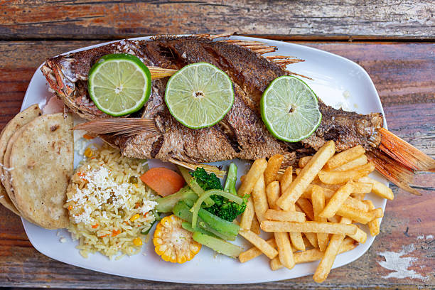 istock A fried whole fish garnished with lime slices and accompanied by various side dishes on a white plate over a wooden table. 2241338262