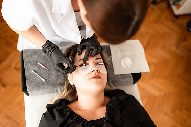 Woman receiving eyebrow tint and eyelash curling treatment from cosmetologist in modern salon.