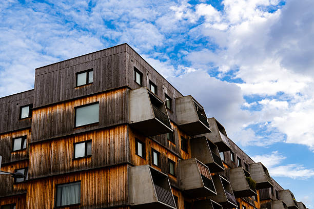 This image showcases a contemporary residential building combining raw concrete balconies with a warm wooden facade. The angular, box-like balconies project outward in rhythmic repetition, creating a dynamic and sculptural appearance. The contrast between natural wood textures and industrial concrete elements highlights a balance between modern design and organic materiality. Set beneath a partly cloudy blue sky, the composition emphasizes depth, geometry, and the innovative aesthetics of sustainable urban housing.