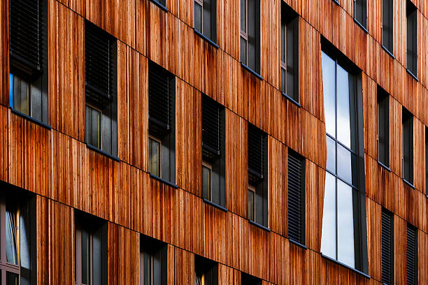 This image depicts a modern residential complex featuring a harmonious combination of wood and concrete materials. The design emphasizes open balconies, horizontal lines, and natural textures that blend with the surrounding environment. The warm wooden panels contrast beautifully with the neutral tones of concrete, while the courtyard layout and stairway create a sense of openness and community. The architecture reflects contemporary urban living focused on comfort, sustainability, and aesthetic balance.
