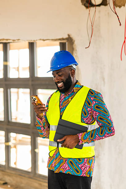 istock Happy African construction engineer in blue hardhat and safety vest using smartphone on building site 2241358549