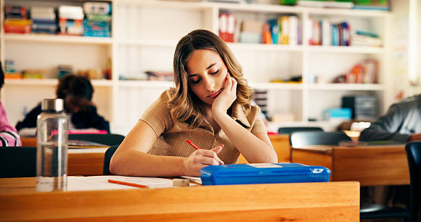 istock Writing, education and student bored in classroom for college admission, thinking or quiz. Aptitude assessment, test and academy with tired girl learning in high school for lesson or scholarship exam 2241360571