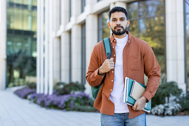 istock A confident young man with a backpack poses in front of a building, holding books. 2241365332