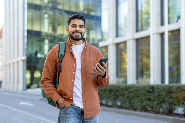 istock Happy man stands outside, smiling, holding a smartphone and wearing a backpack 2241365354