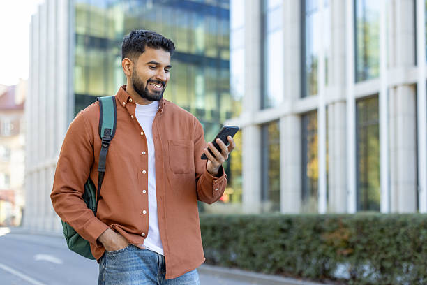 istock A smiling student, outdoors, using his phone while walking in front of a modern building. 2241365371