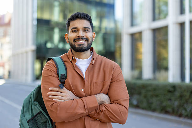 istock A smiling Indian student with a backpack stands confidently outside a modern building, with arms crossed. 2241365376