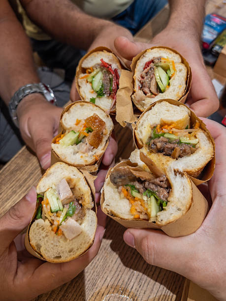 Multiple hands share delicious Vietnamese banh mi sandwiches, showcasing fresh meat, pickled vegetables, and herbs. A vibrant, authentic Asian street food experience shared on a wooden table.