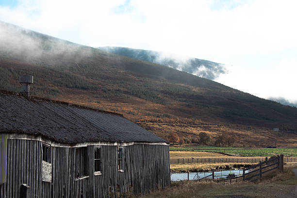 Rustic abandoned wooden building in a scenic rural landscape, with mist rolling over the hills in the Scottish Highlands. The old weathered structure stands beside a river, framed by a fence and surrounded by autumn-colored fields, creating a moody, atmospheric scene of nature and decay.