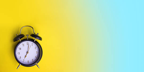 istock Black alarm clock on a yellow and blue empty background. Minimalist composition with copy space and bright contrasting colors. 2241374125