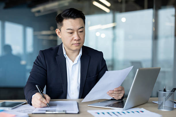 istock Serious young Asian man in a suit working in the office on documents, studying papers and writing down data 2241375083