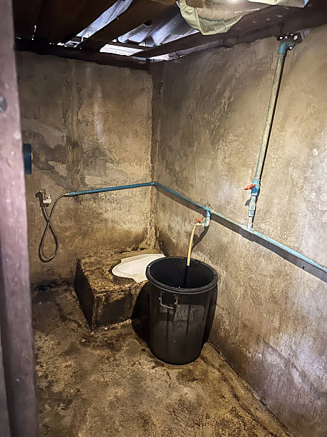A rustic and basic outdoor squat toilet facility featuring exposed plumbing, a water bucket, and rough concrete walls. This image depicts rudimentary sanitation conditions in a rural or developing
