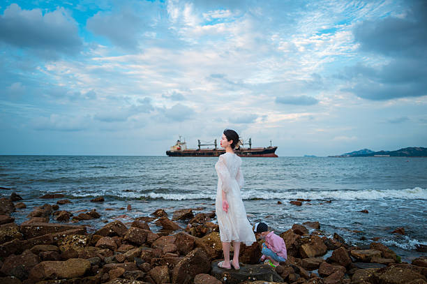 A woman in a white long dress walking towards the seaside, with her back to the camera. In the distance, a large cargo ship is quietly docked on the water, as gentle waves lap against the shore, creating a serene and poetic atmosphere