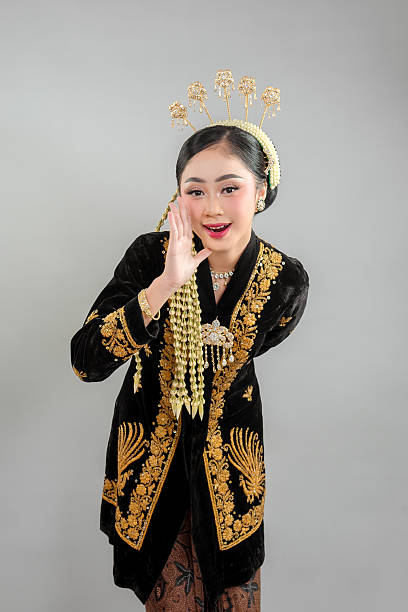 istock Smiling Asian woman in traditional Javanese attire leans forward with a cheerful expression, hand near mouth, as if happily sharing a secret or greeting someone warmly. 2241384123