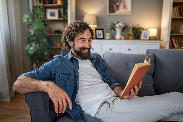 istock Happy man relaxing on sofa reading book at home 2241387169