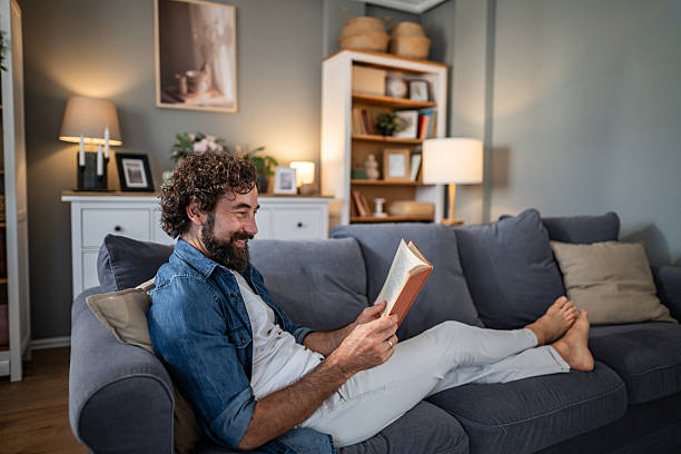 istock Man relaxing on sofa reading a book barefoot in living room 2241387181