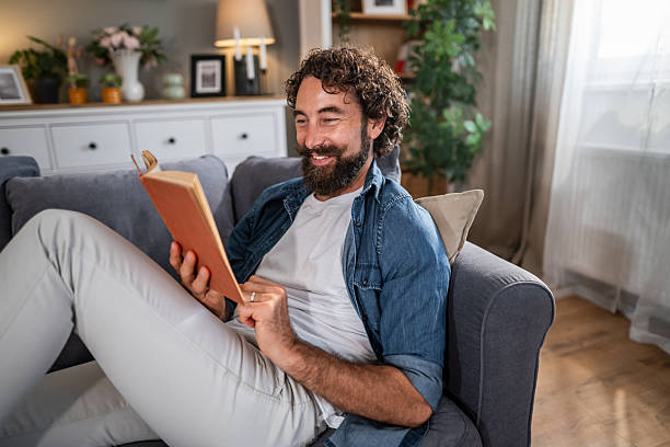 istock Man relaxing on sofa reading book and smiling 2241387269
