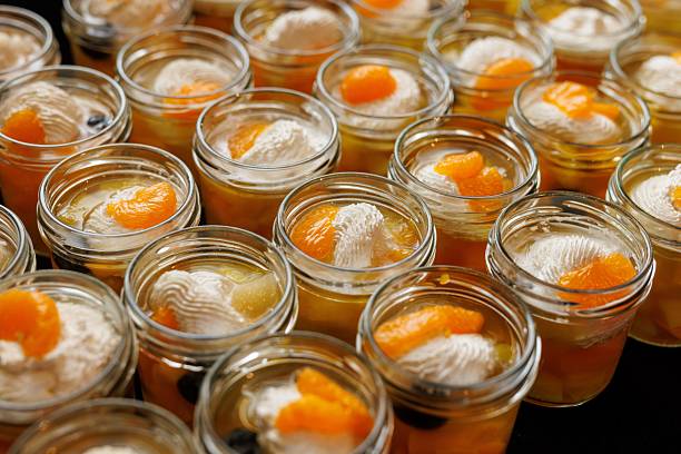 Multiple glass jars filled with layered dessert, featuring mandarin slices and whipped cream. Ideal for catering, parties, and dessert-themed visuals.
